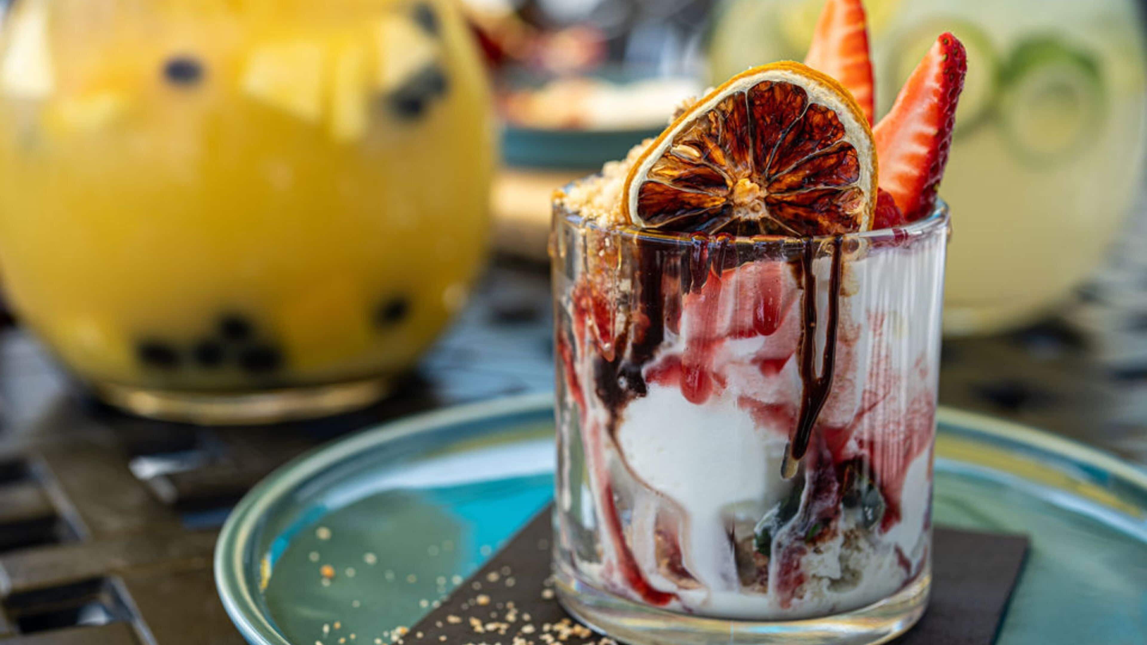 Bebida postre en un vaso con una rodaja de naranja deshidratada y una fresa, servida sobre un plato de vidrio con borde azul.