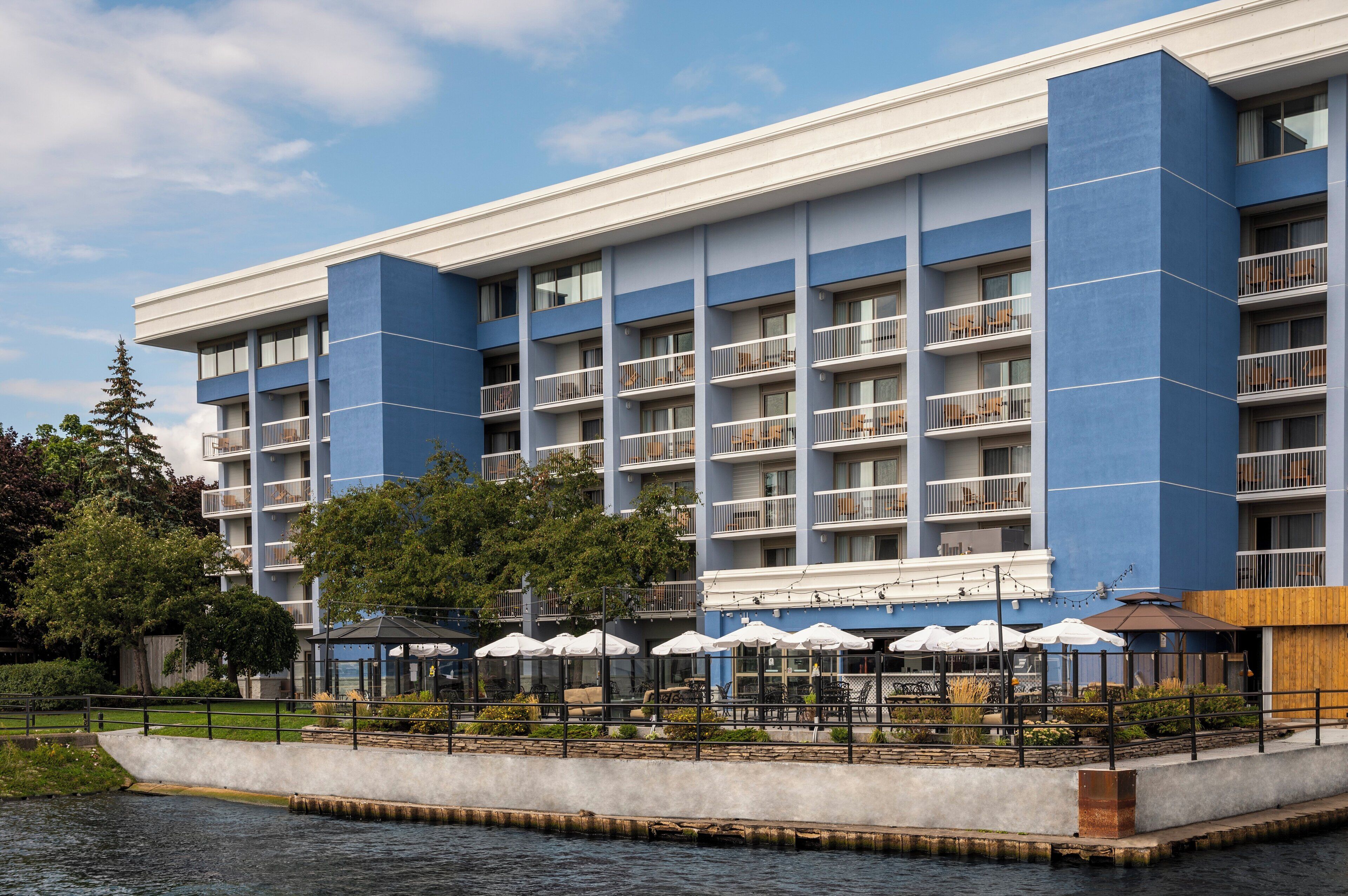 A modern hotel with multiple balconies, a waterfront patio with umbrellas, and a pool area.