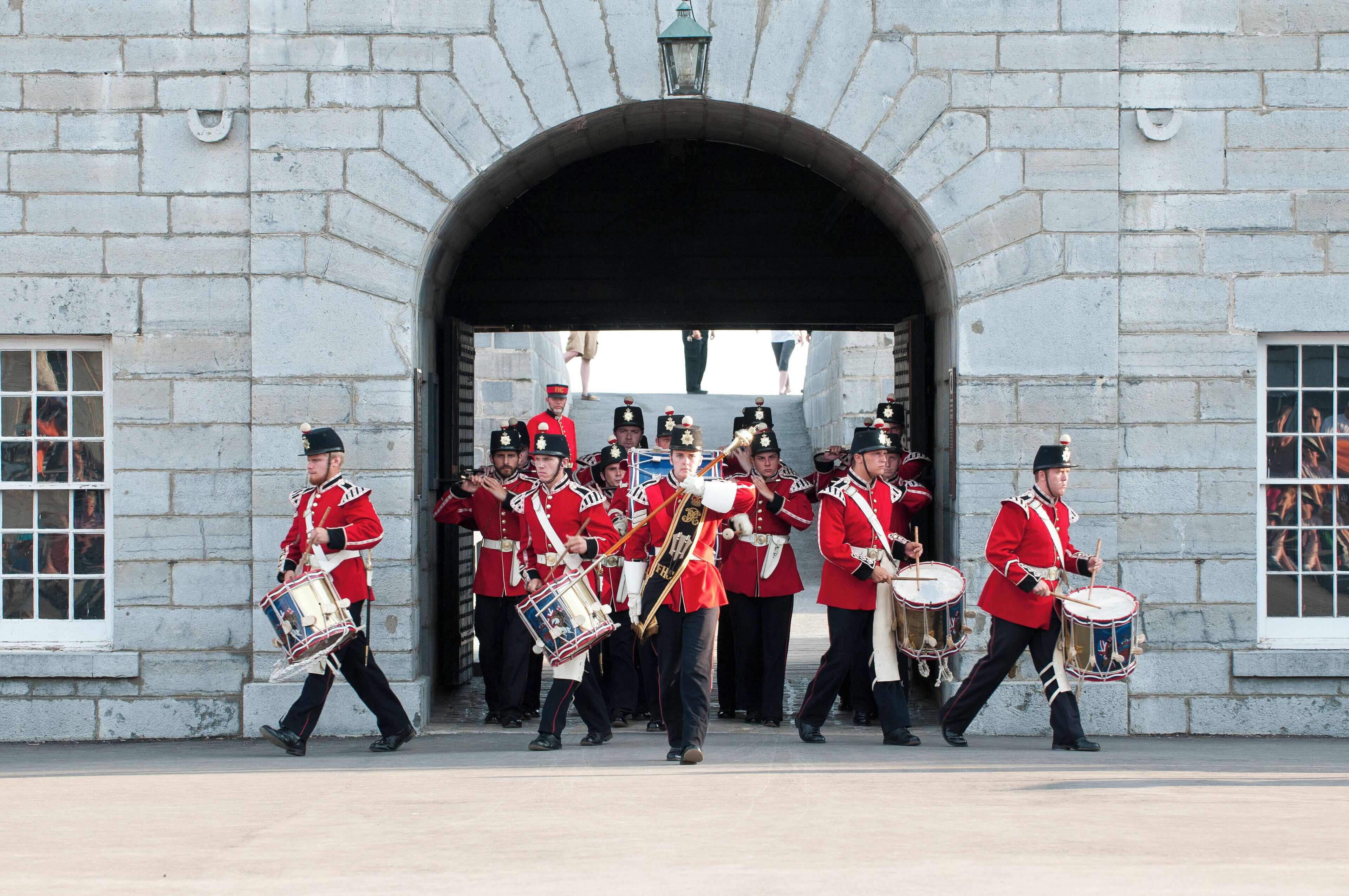 A group of uniformed guards marching in formation, carrying drums, in front of a stone building with an arched entrance.