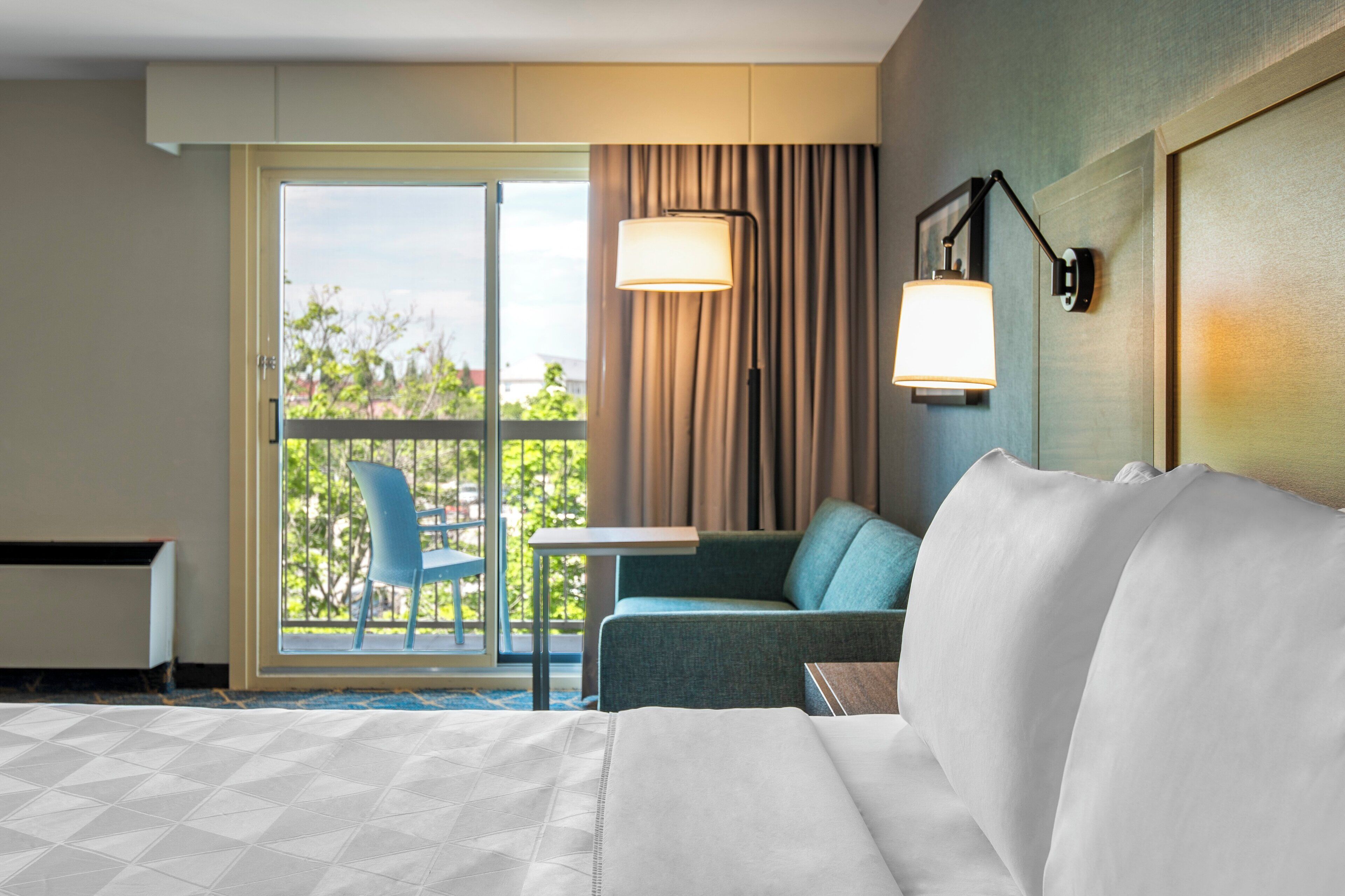 A hotel room with a bed, a sofa, a table, and a chair. There is a view of greenery outside the window.