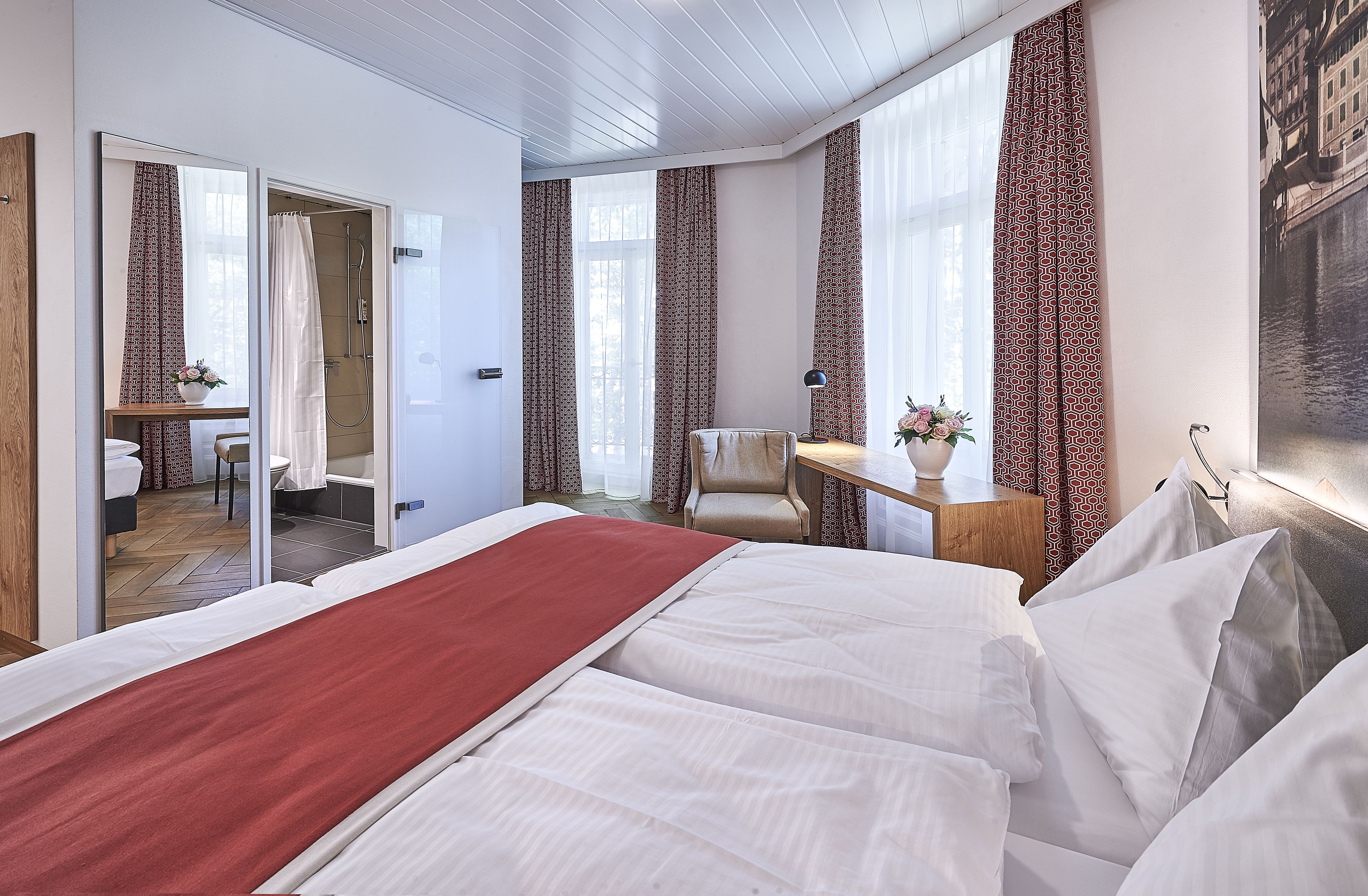 Ein Hotelzimmer mit einem großen Bett, einem Schreibtisch mit Stuhl, einem kleinen Badezimmer und Blick auf ein Gebäude durch das Fenster.