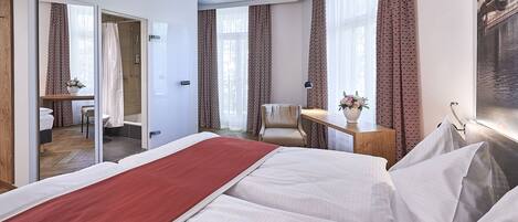 A hotel room with a large bed, a desk with a chair, a small bathroom, and a view of a building through the window.