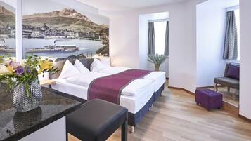 Una habitación de hotel moderna con una cama, una mesita con un florero, una silla y vistas a un paisaje urbano a través de una ventana.