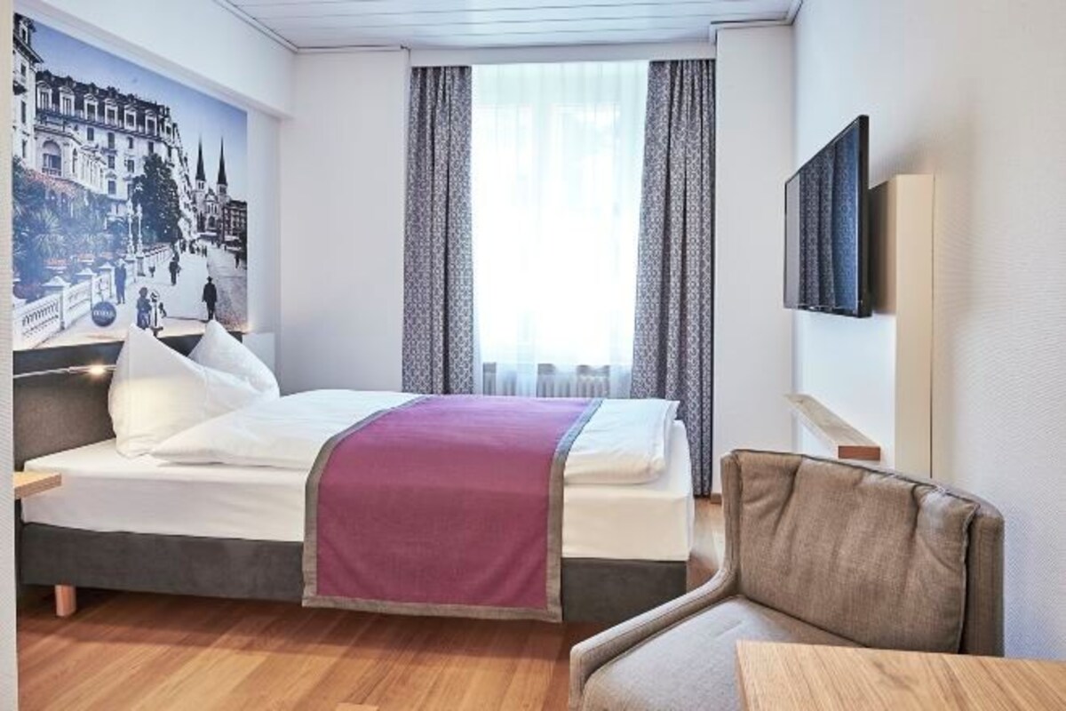 A hotel room with a bed, a chair, a TV, a window with curtains, and a mural on the wall.