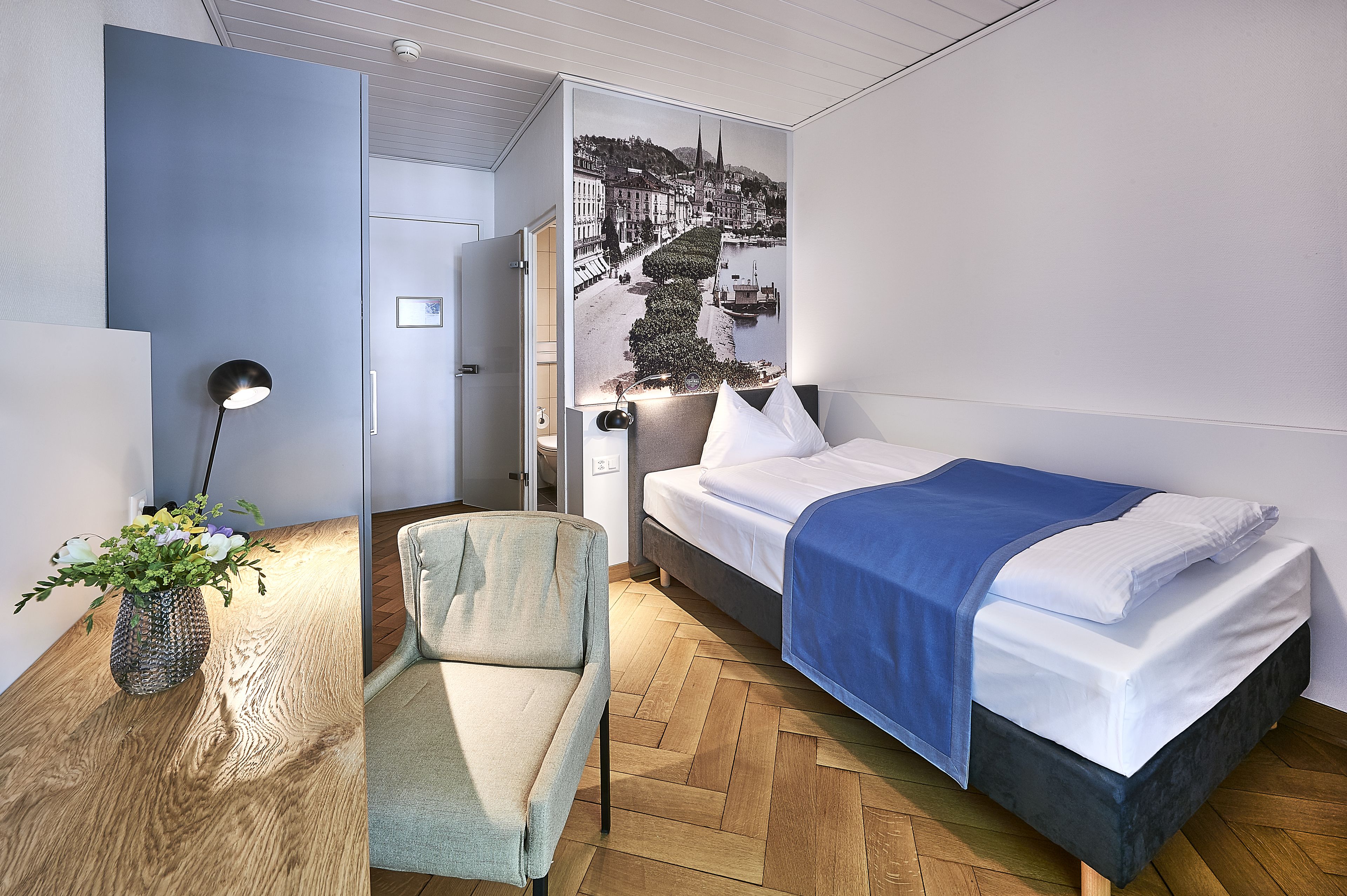Una habitación de hotel moderna con una cama, una silla, un escritorio con lámpara y un mural grande en la pared.