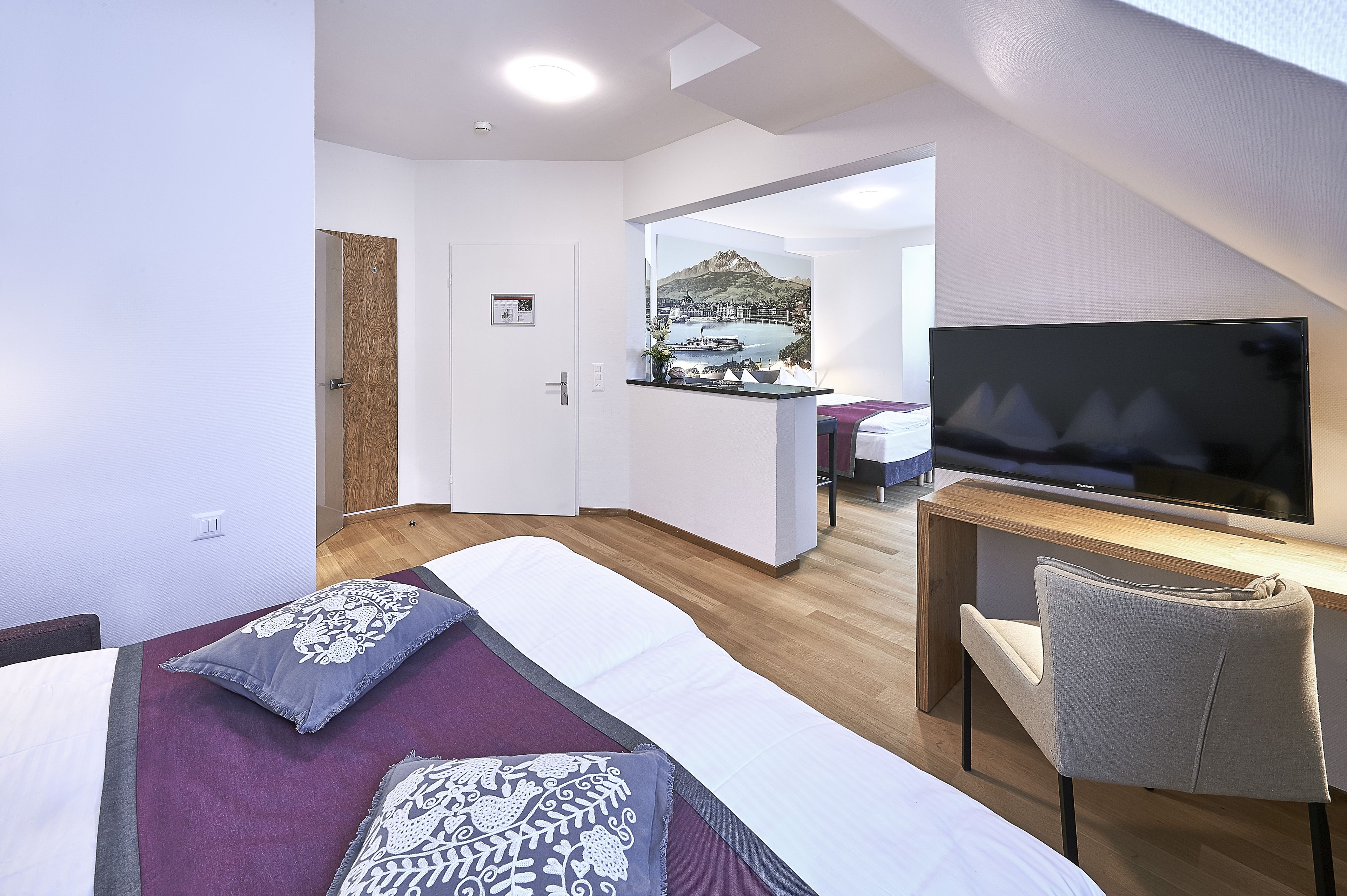A modern hotel room with a bed, a desk, a TV, and a view of a mountainous landscape.