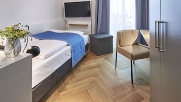 A modern hotel room with a bed, a TV, a small table with a vase, a chair, and a herringbone wooden floor.