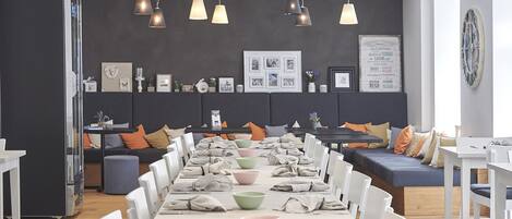 Un comedor moderno con una mesa larga dispuesta para una comida, un sofá con cojines e iluminación colgante.