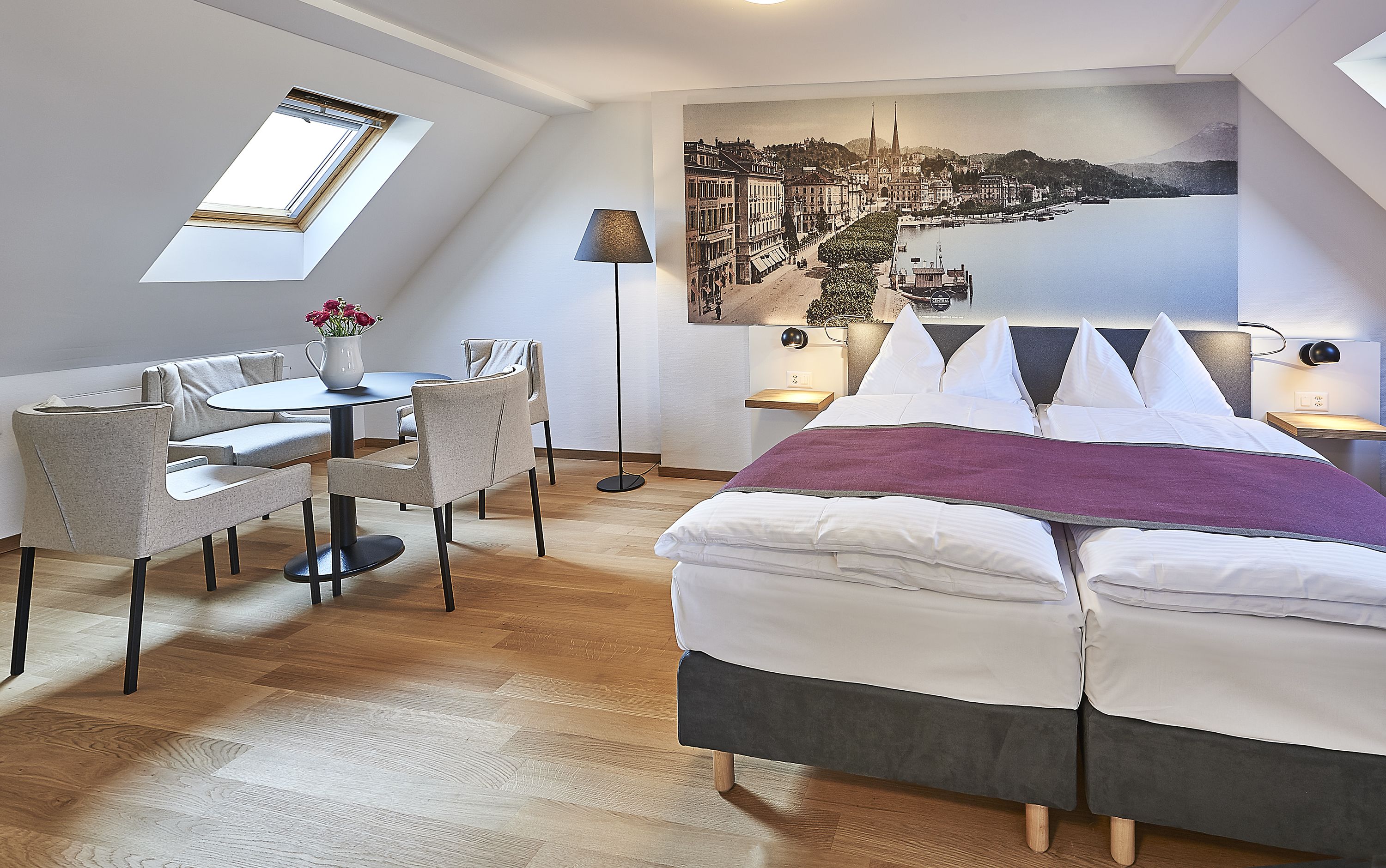 Una habitación de hotel moderna con cama, una mesa de comedor con sillas, un pequeño balcón y un cuadro de paisaje urbano en la pared.