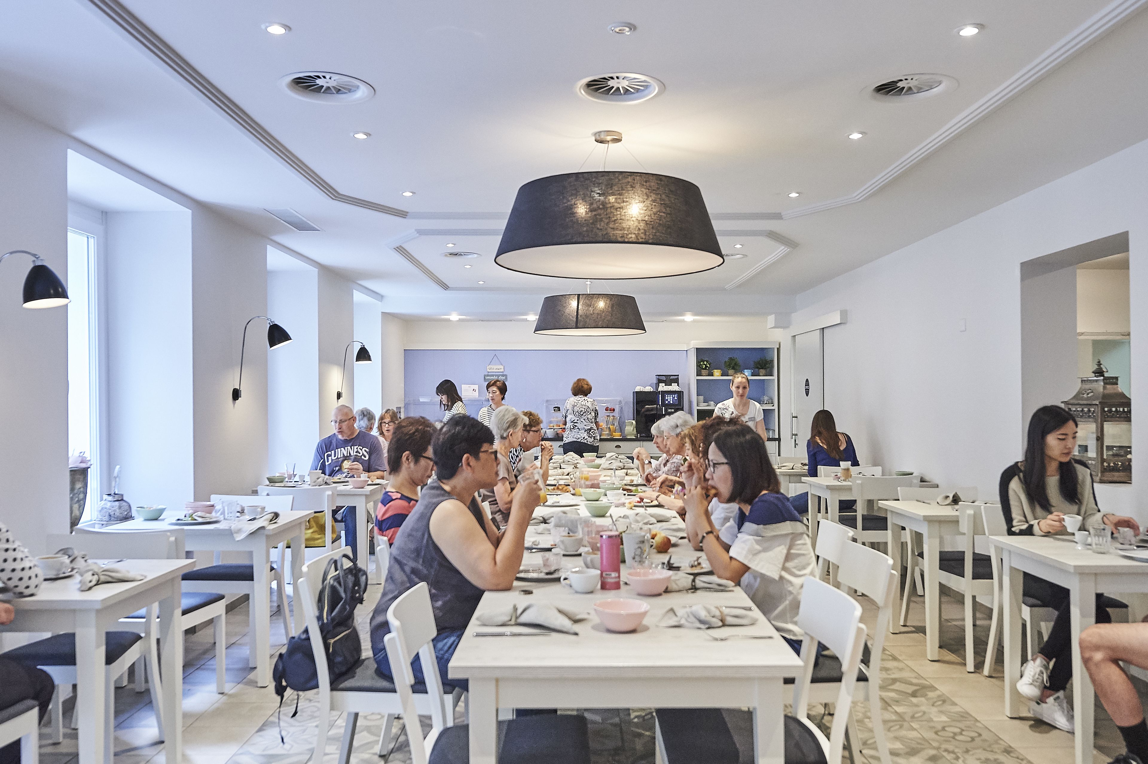 Un espace repas moderne avec des tables et des chaises blanches, un grand luminaire suspendu noir et une vue sur la cuisine en arrière-plan.