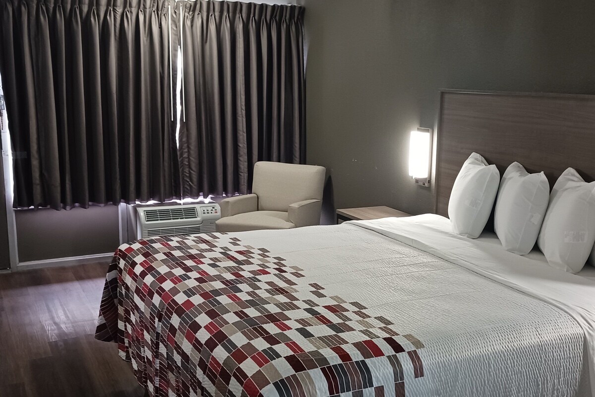 A hotel room with a large bed, a chair, a bedside table, and a window with curtains.