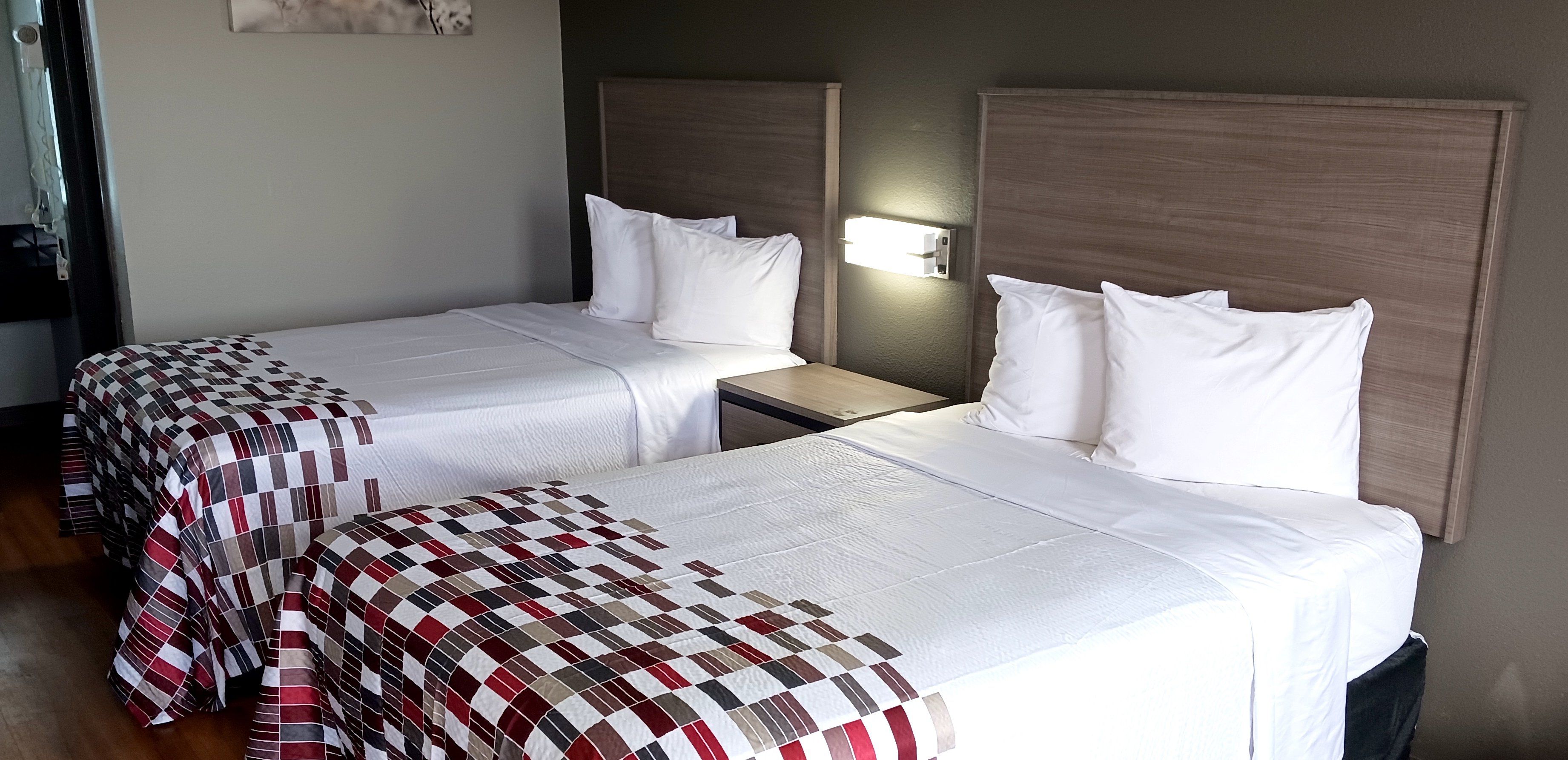 A hotel room with two single beds, each with a white bedspread and pillows. There is a nightstand between the beds with a lamp. The room has a wooden headboard and a wall-mounted light fixture.