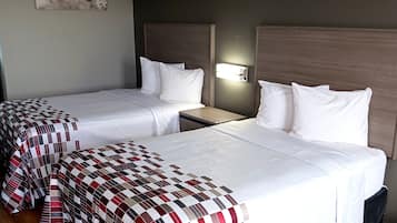 A hotel room with two single beds, each with a white bedspread and pillows. There is a nightstand between the beds with a lamp. The room has a wooden headboard and a wall-mounted light fixture.