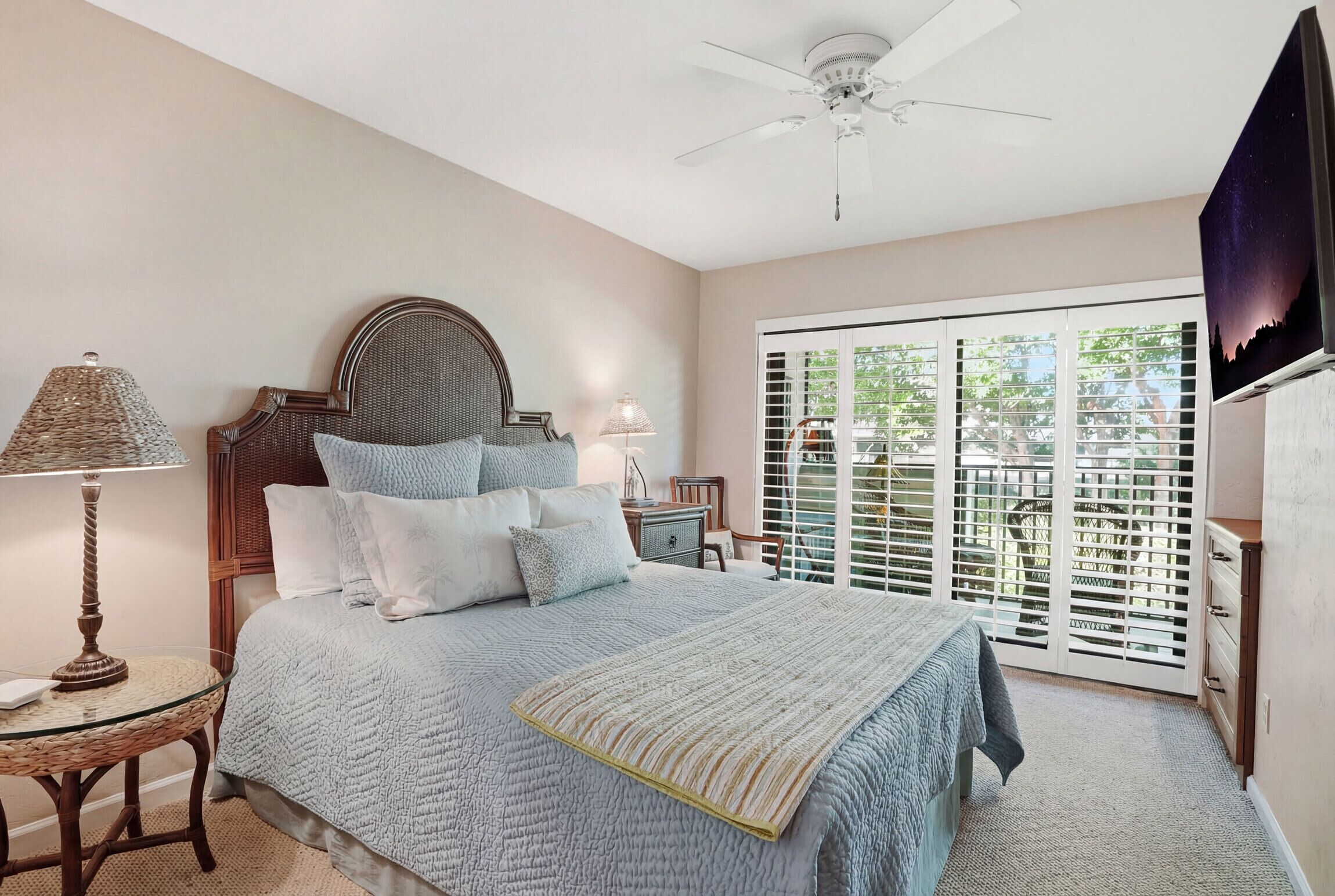 A bedroom with a bed, a bedside table, a lamp, a ceiling fan, and a view through the window.