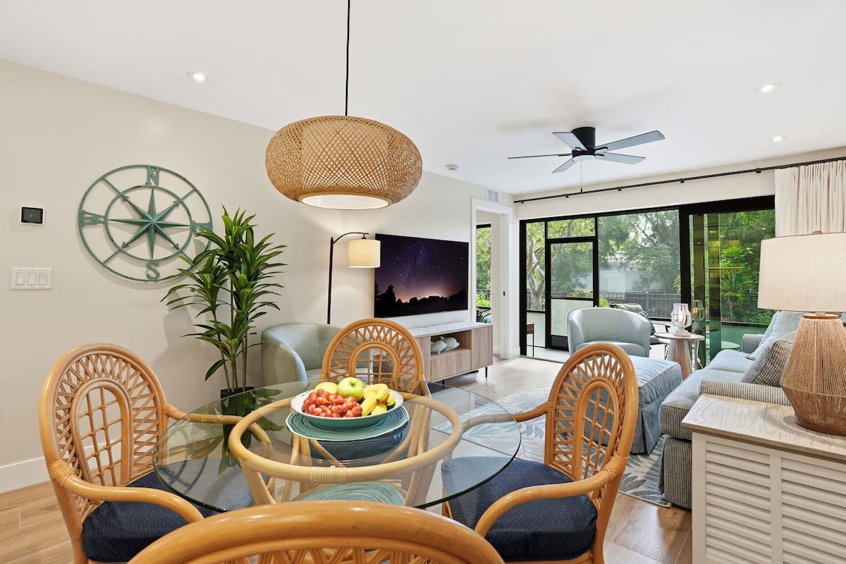 A modern living room with a glass dining table, wicker chairs, a TV, and a large window.