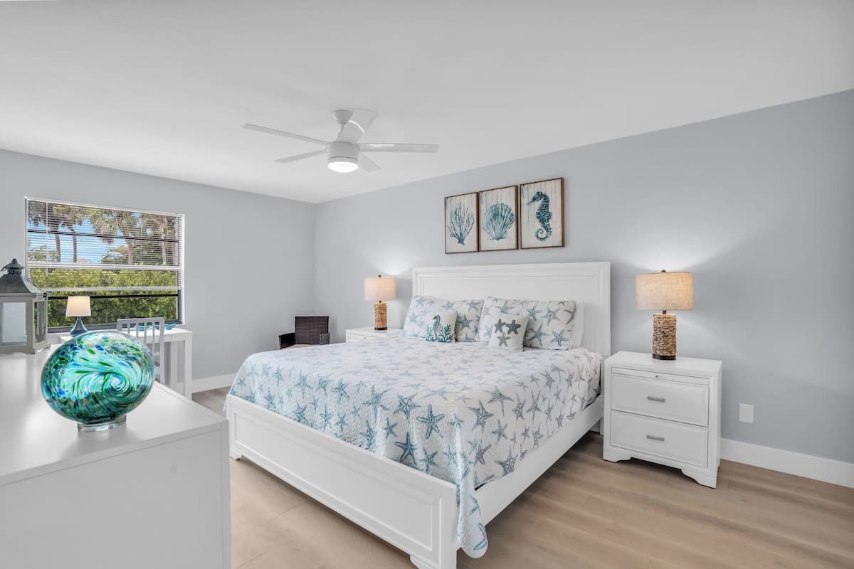 A bedroom with a bed, nightstands, a lamp, a ceiling fan, and artwork on the wall.