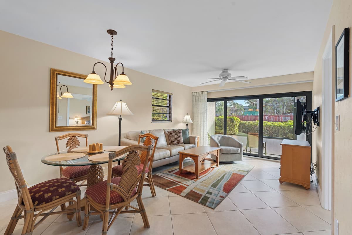 A living room with a dining table, chairs, a sofa, a ceiling fan, and a large window.