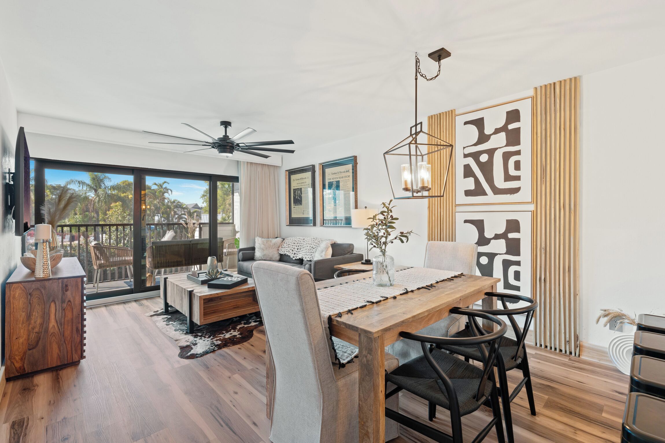 A modern living room with a dining area, large windows, ceiling fan, and decorative wall panels.