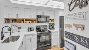 A modern kitchen with white cabinets, a black stove, and a refrigerator. There are signs on the wall that read "Coffee Bar" and "Fresh Produce." - Park Shore Resort (Naples)