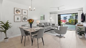 A modern living room with a dining area, a grey sofa, a ceiling fan, and a large window with a view of buildings and greenery. - Park Shore Resort (Naples)
