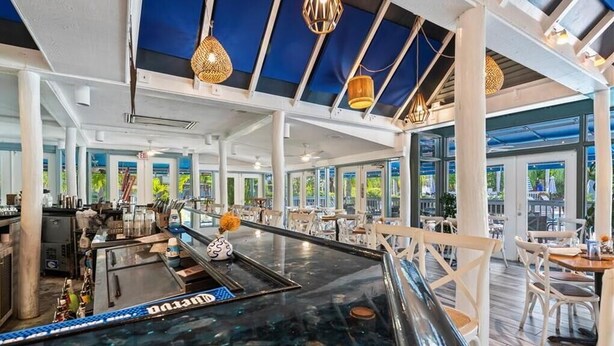 An indoor pool area with a glass floor, seating around the pool, and a bar area with a blue ceiling and hanging lights. - Park Shore Resort (Naples)