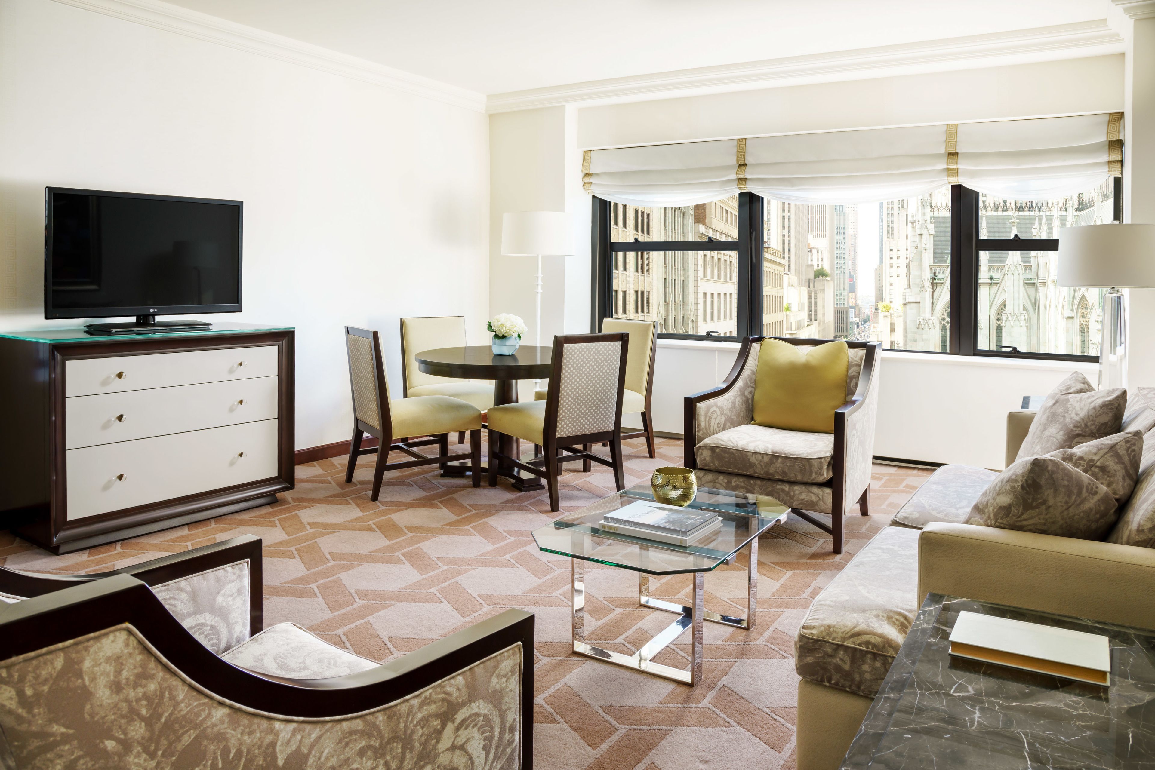 A hotel room with a flat-screen TV, a dining table with chairs, a sofa, and a large window with a city view.
