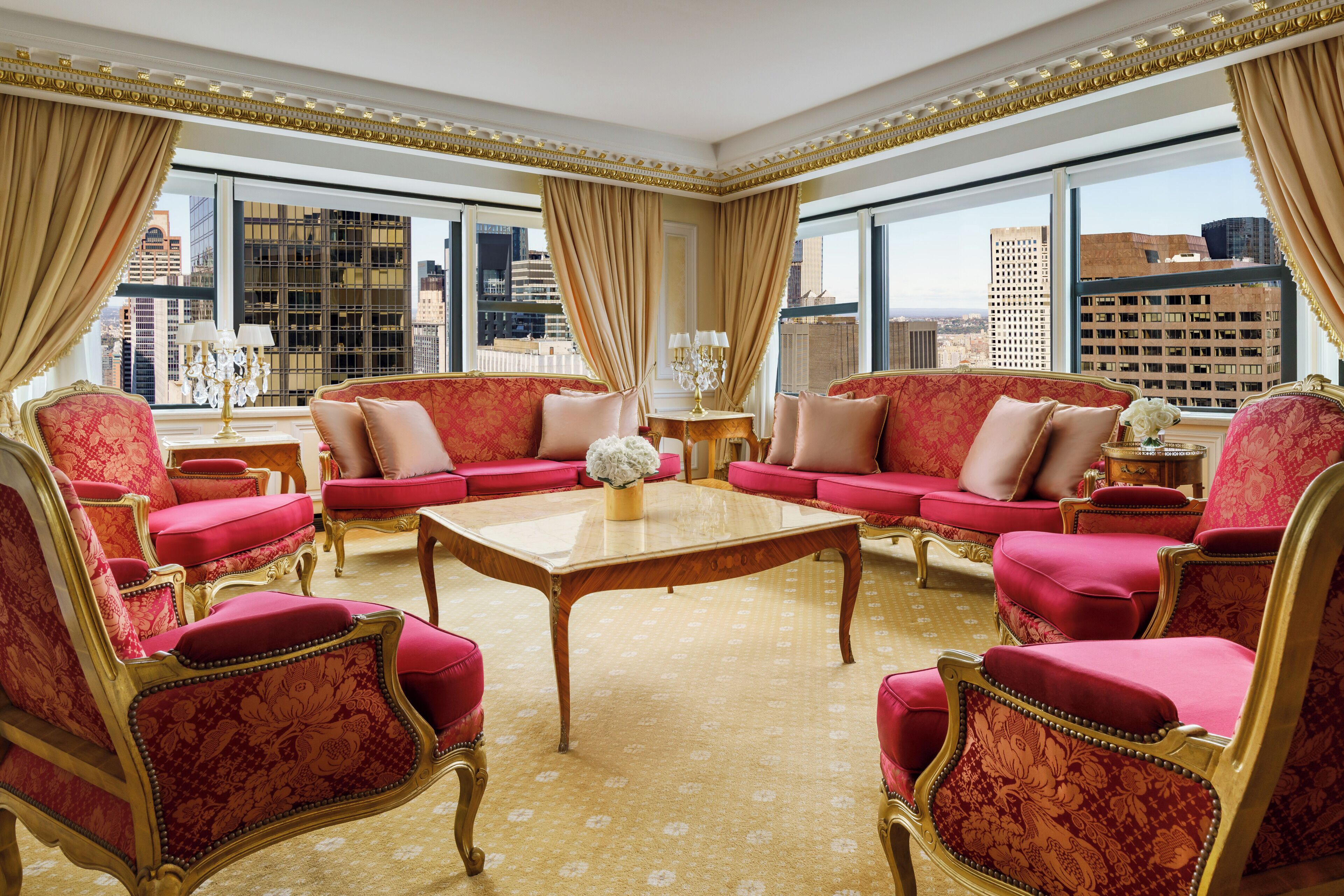 Una habitación lujosa con muebles tapizados en rojo, una mesa de centro de madera y grandes ventanales con vistas a la ciudad.
