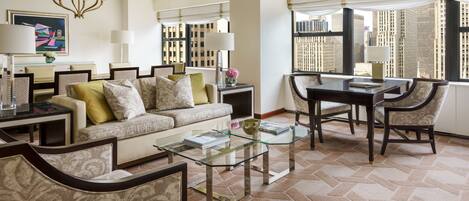 A modern living room with a glass coffee table, a sofa with patterned cushions, a dining table with chairs, and large windows with a city view.