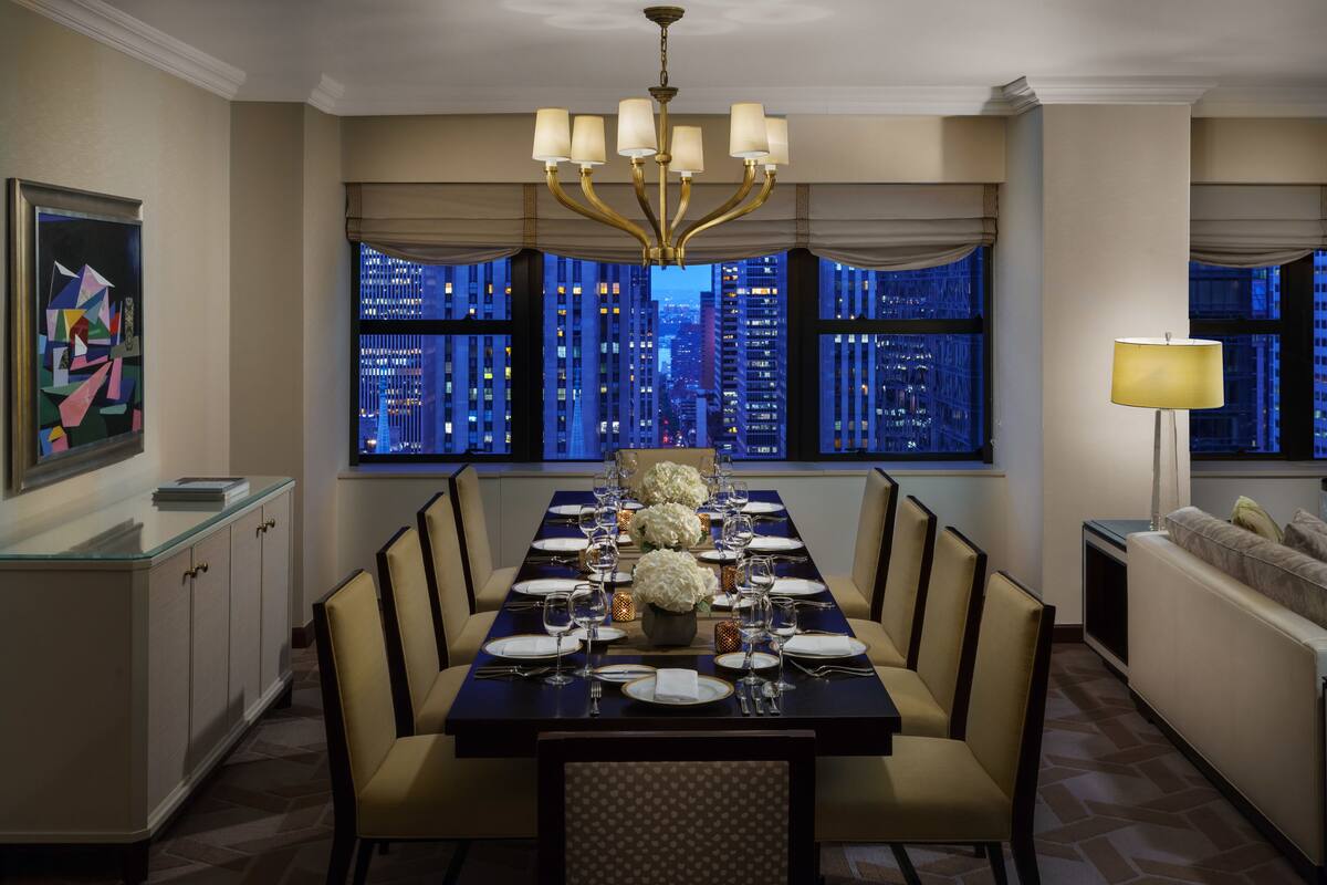 Een eetkamer met een lange tafel voor zes personen, grote ramen met uitzicht over de stad, een kroonluchter en een schilderij aan de muur.