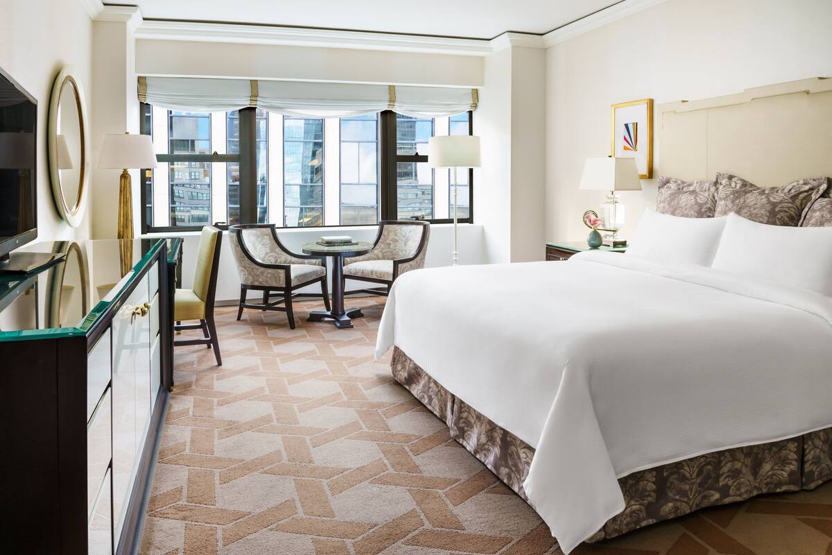 Een hotelkamer met een groot bed, een bureau met twee stoelen, een televisie en uitzicht op de stad door grote ramen.