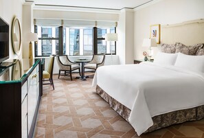 A hotel room with a large bed, a desk with two chairs, a television, and a view of the city through large windows.