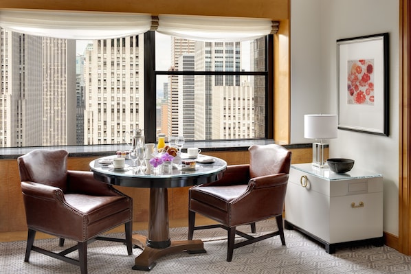 Quarto de hotel com vista para a cidade, uma mesa de jantar redonda com cadeiras, uma mesinha de cabeceira com abajur e uma obra de arte emoldurada na parede.