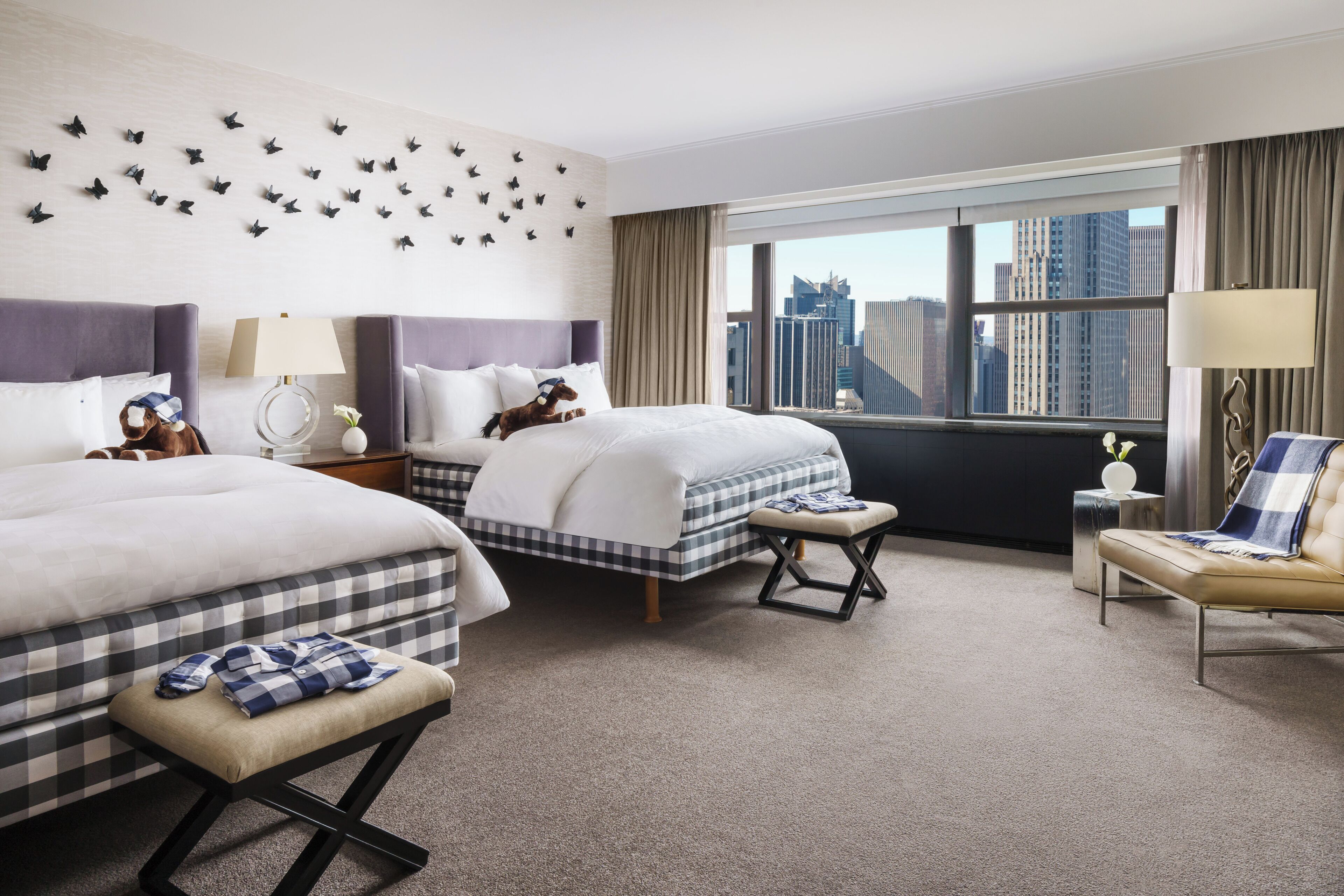 Habitación de hotel con dos camas, un banco, una mesita, una lámpara y vistas a la ciudad.