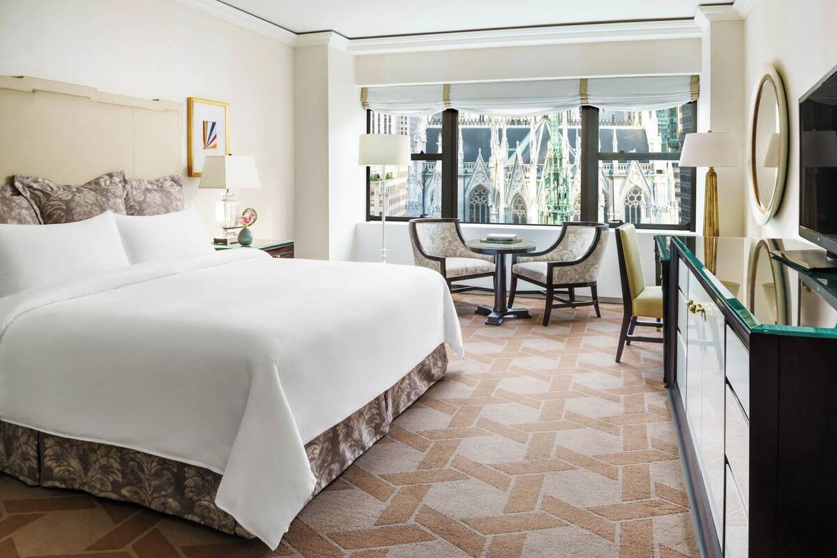 Een hotelkamer met een groot bed, een eettafel met stoelen, uitzicht op een gebouw met bijzondere architectuur en een televisie op een kast.