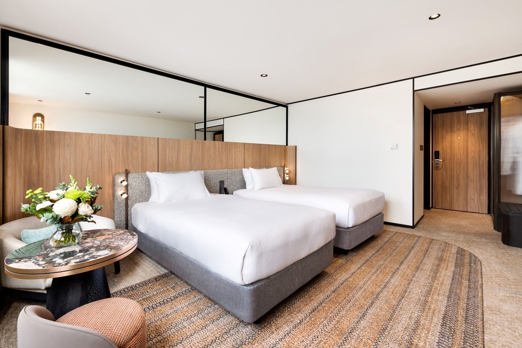 A modern hotel room with two beds, a round table with a vase of flowers, and a large mirror.