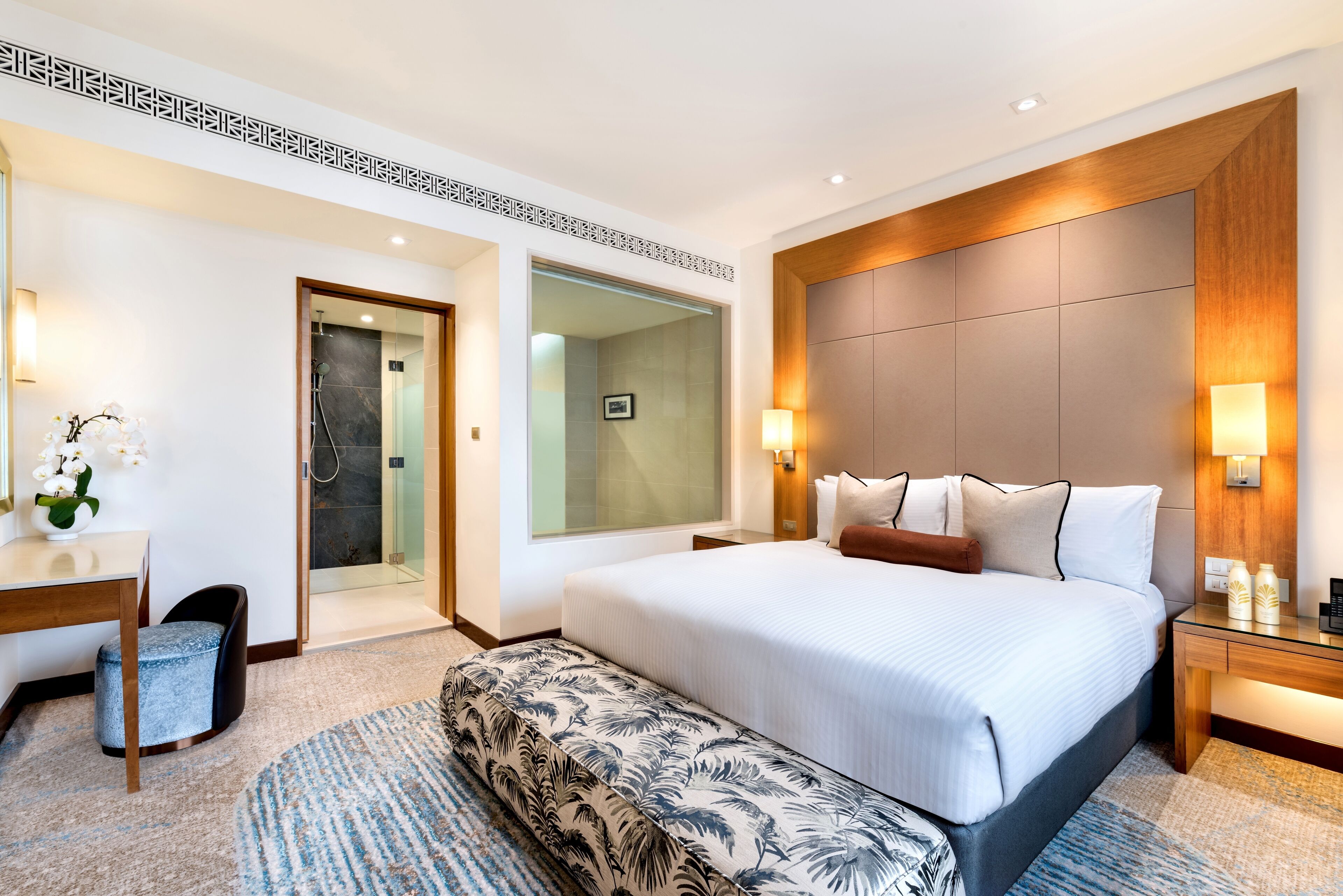 Habitación de hotel con una cama grande, un escritorio, un banco, una ducha y un espejo.