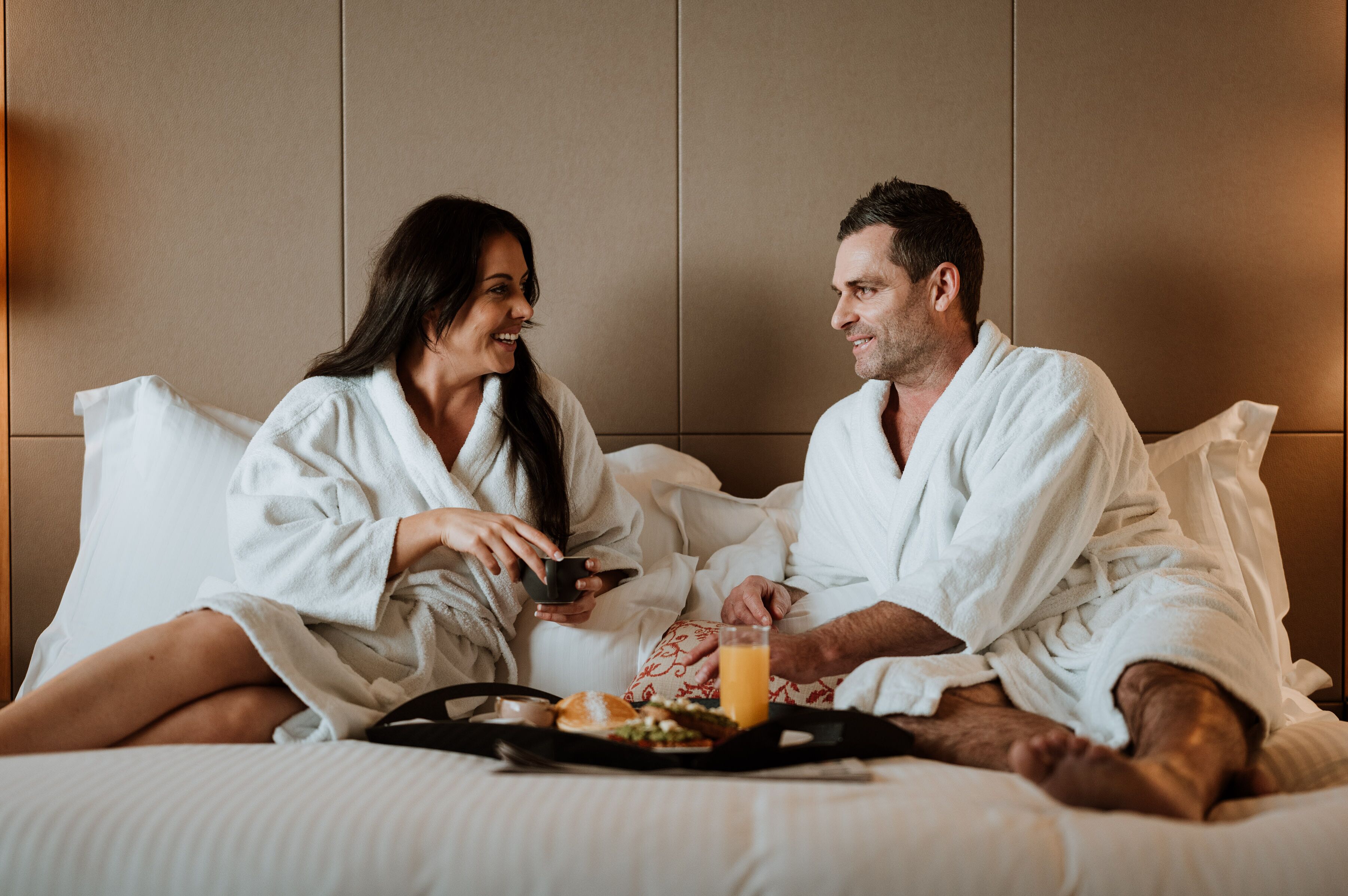 A couple in white bathrobes sitting on a bed with a tray of breakfast including toast, a bowl, and a glass of orange juice.
