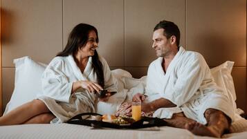 A couple in white bathrobes sitting on a bed with a tray of breakfast including toast, a bowl, and a glass of orange juice.