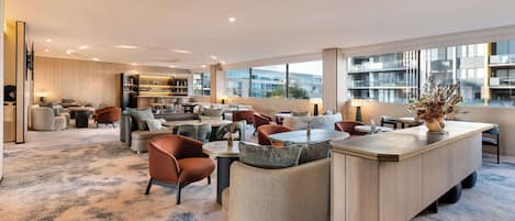 A modern hotel lobby with a spacious seating area, a bar, and a large window offering a view of the city.