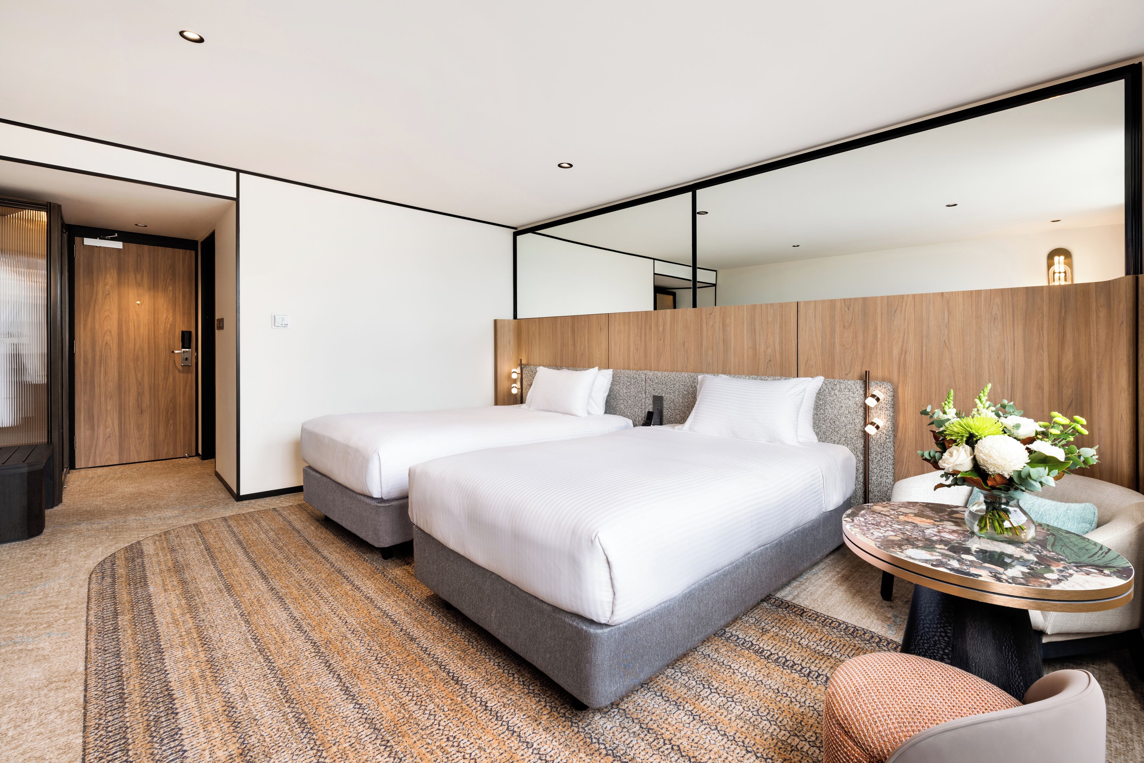 A modern hotel room with two beds, a round table with a vase of flowers, and a large mirror on the wall.