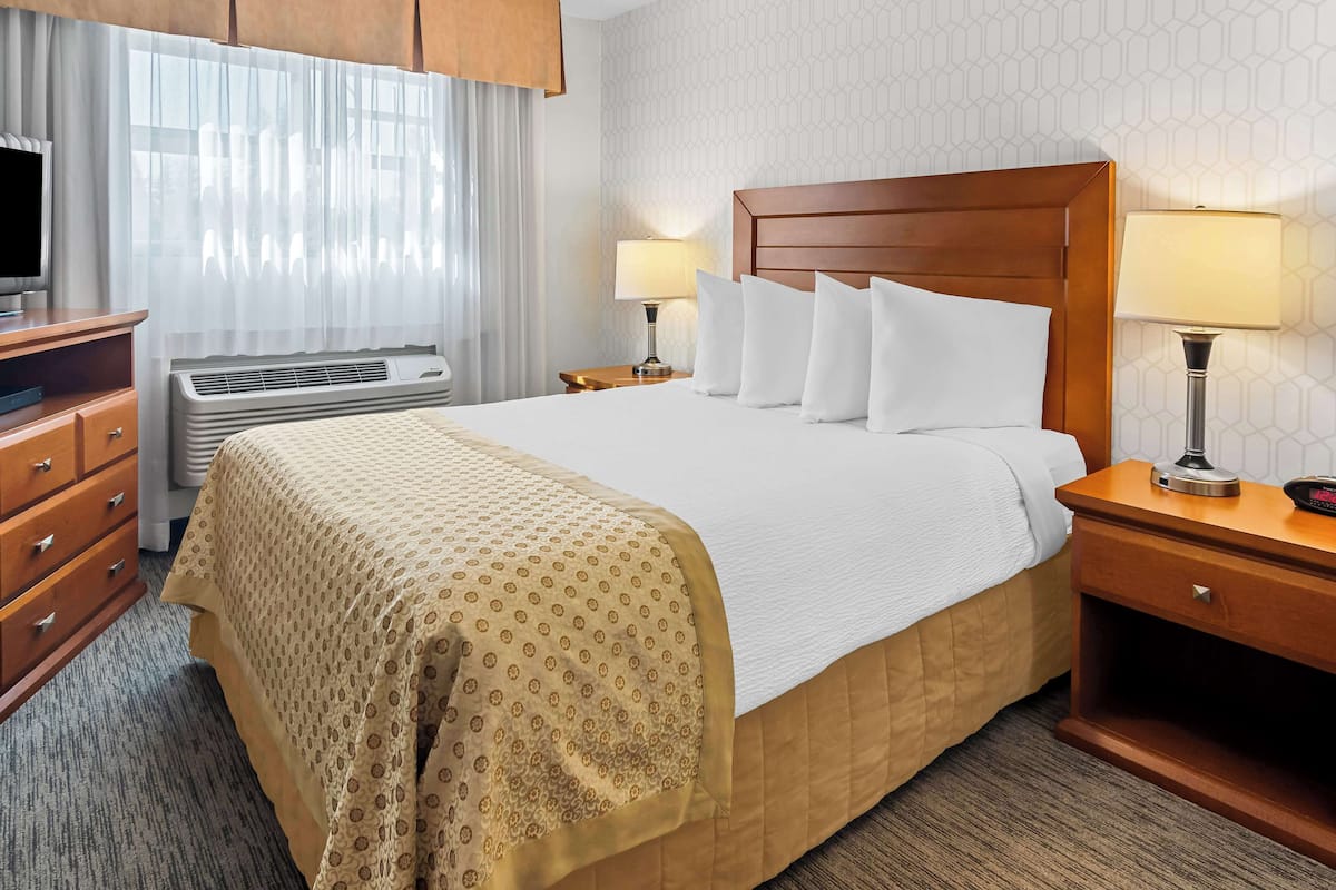 A hotel room with a large bed, two bedside tables with lamps, a TV, a dresser, and an air conditioning unit.