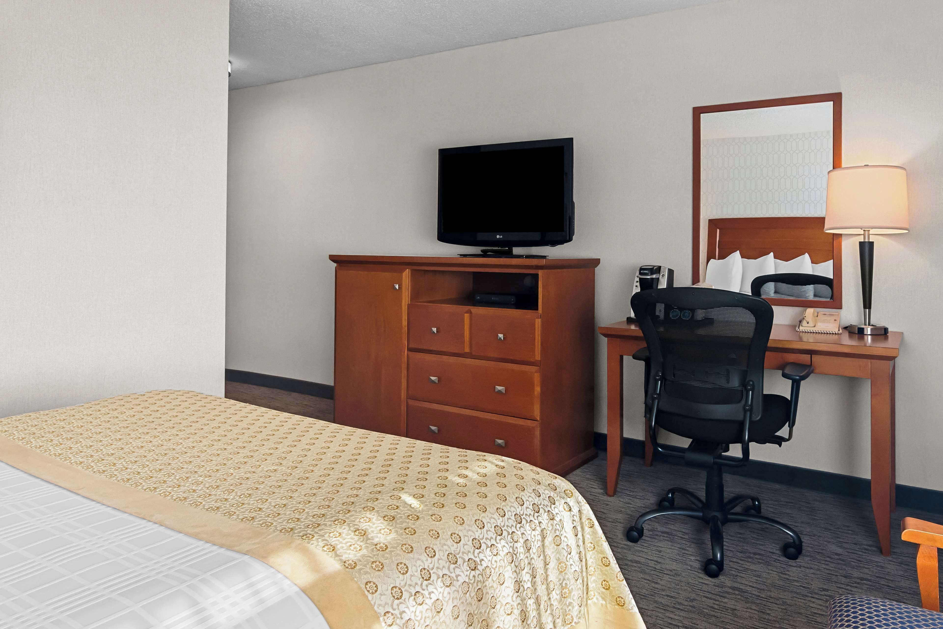 Habitación de hotel con cama, cómoda de madera, escritorio con computadora y silla, televisor sobre la cómoda y una lámpara en el escritorio.