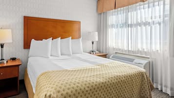 A hotel room with a large bed, a wooden headboard, two bedside tables with lamps, a window with sheer curtains, and an air conditioning unit.