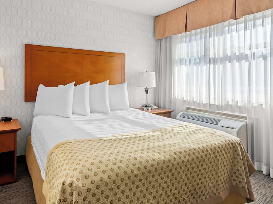 A hotel room with a large bed, a wooden headboard, two bedside tables with lamps, a window with sheer curtains, and an air conditioning unit.