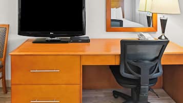 A hotel room with a wooden desk, a black office chair, a flat-screen TV, a telephone, and a mirror reflecting a bed with white pillows.
