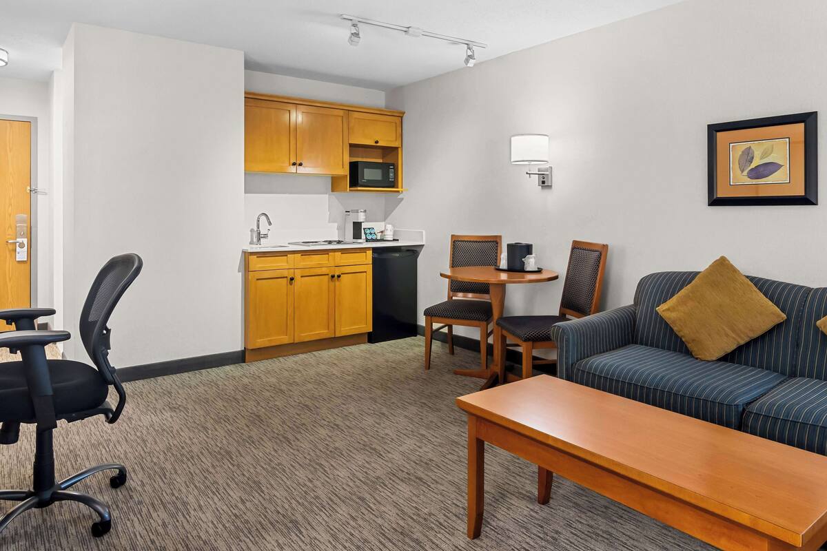 A compact hotel room with a kitchenette, a sofa, a dining table, and a single office chair.