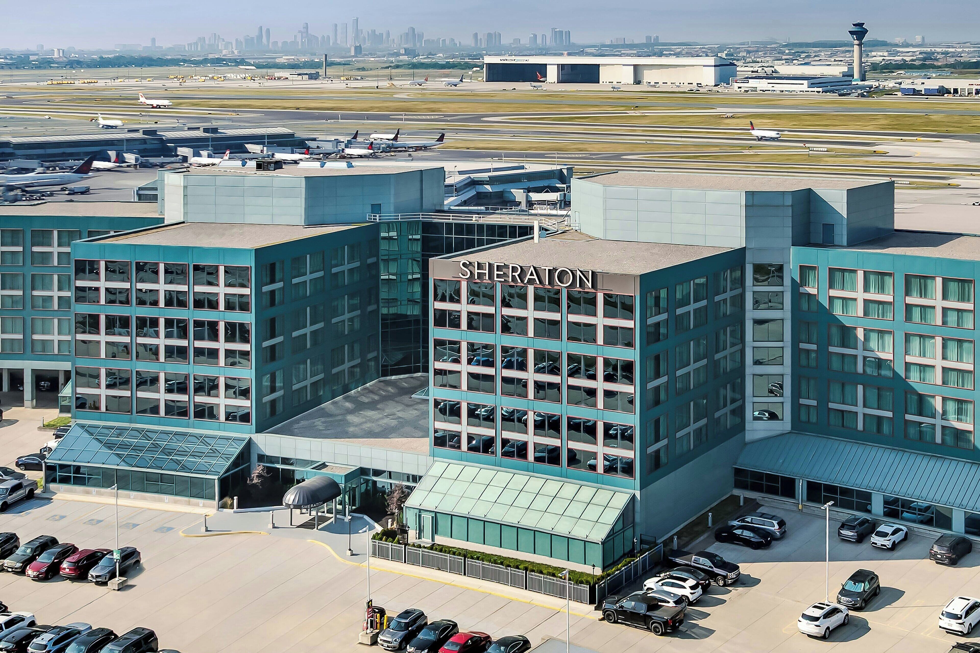 Un hotel Sheraton con fachada de vidrio moderno, varios niveles de estacionamiento y vista al aeropuerto de fondo.