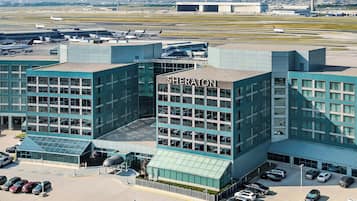 Un hotel Sheraton con fachada de vidrio moderno, varios niveles de estacionamiento y vista al aeropuerto de fondo.
