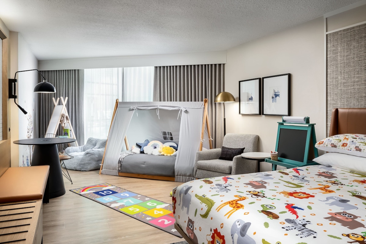 Una habitación de hotel moderna con una cama grande, una zona de descanso acogedora, una tienda de juegos y un pequeño escritorio con lámpara.