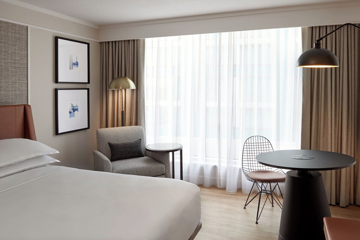 Una habitación de hotel con una cama grande, una mesita redonda pequeña, una silla y una lámpara.